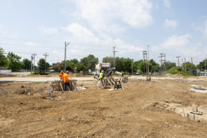 A volumetric concrete mixer places concrete into a wooden pedestal form in the background as two concrete finishers trowel another form in the foreground of a construction site in Tulsa, OK.